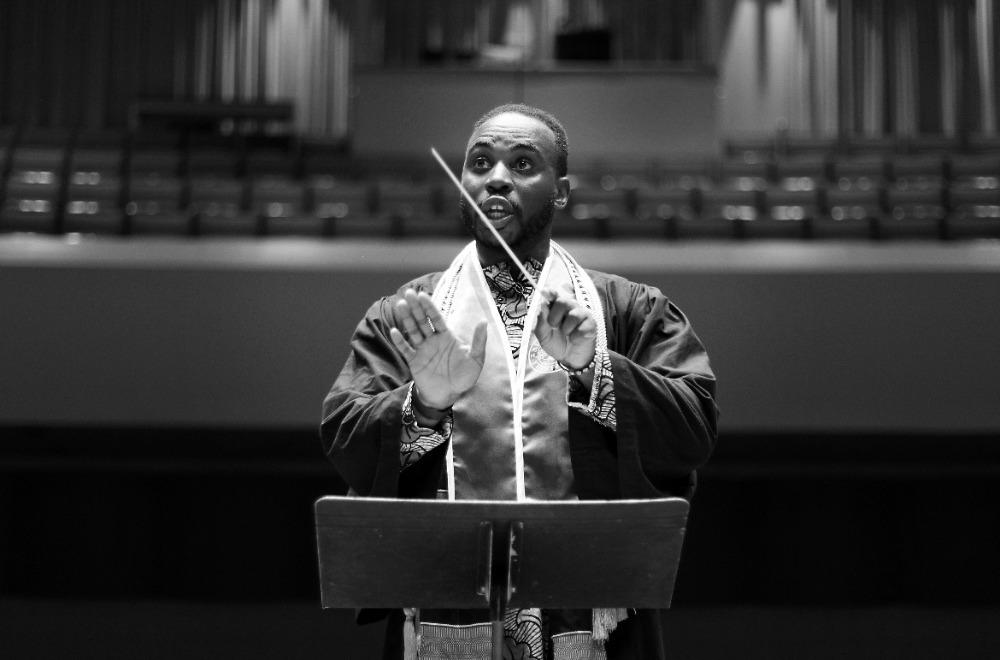 A black 和 white photo of a Black male student conductor wearing graduation regalia conducting an ensemble.